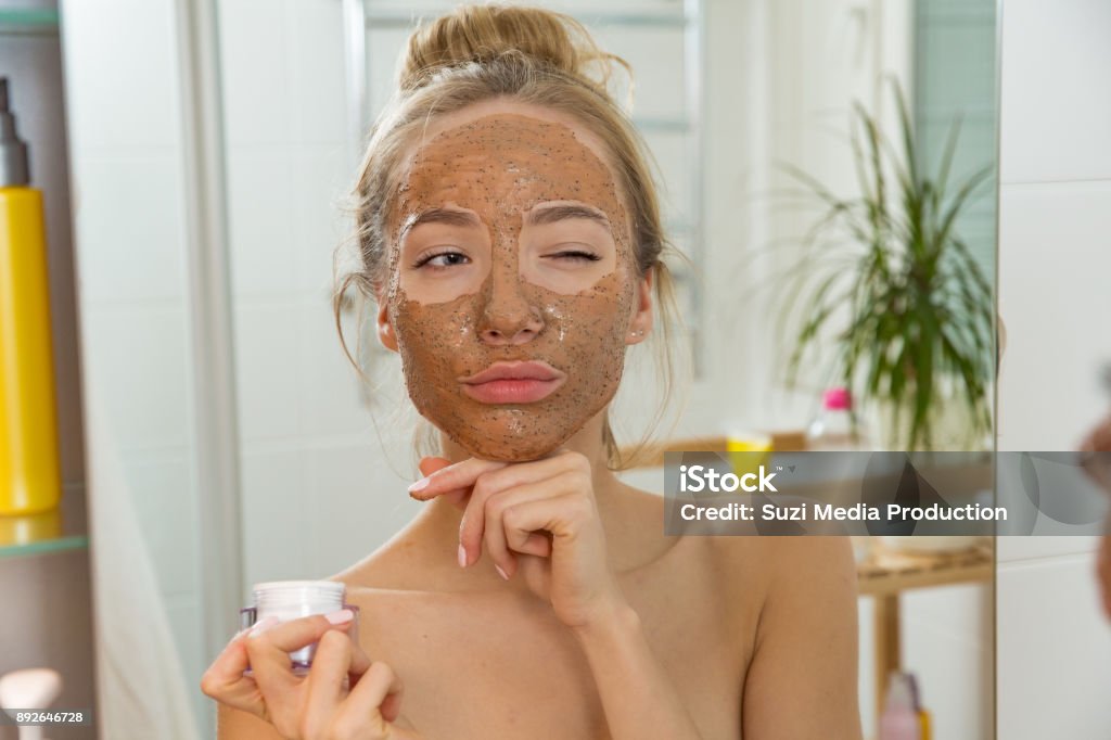 Young beautiful girl applying facial scrub mask on skin. Young beautiful girl applying facial scrub mask on skin. Looking in the mirror in bathroom, Wrapped in a towel, having fun. Exfoliation Stock Photo
