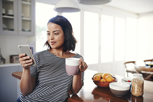 Shot of an attractive young woman using her cellphone while drinking coffee at home