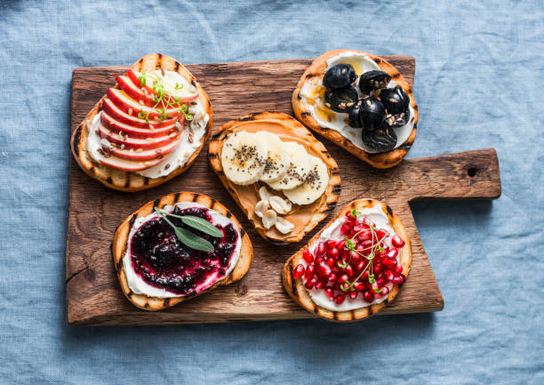 variety grilled bread dessert small plates sandwiches with cream cheese and apple, pomegranate, jam, grapes, peanut butter, banana, flax seed, chia, nuts on a rustic cutting board on blue background, top view. flat lay. delicious breakfast or snack - crostini imagens e fotografias de stock