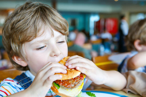istock Cute healthy preschool boy eats hamburger sitting in school canteen 892780408
