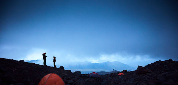Camp in the mountains with 2 people trying to call with a satellite phone, tents in the foreground and a threatening sky in the background