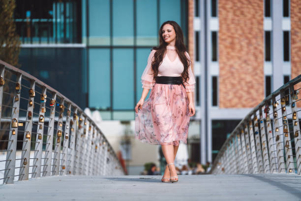 A beautiful teenage girl wearing pink fluffy shirt and a long pink skirt, standing on a bridge full of love locks, smiling, walking around, feeling happy and cheerful, having long brown and curly hair, wearing beige heels and enjoying in the nature full of sun and autumn leaves.