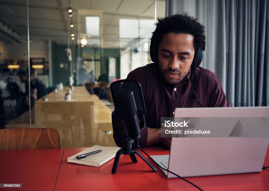 Hosting an online talk show Shot of a young man recording a podcast on a laptop in a modern office Journalist Stock Photo