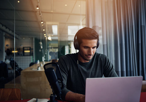 Shot of a young man recording a podcast on a laptop in a modern office