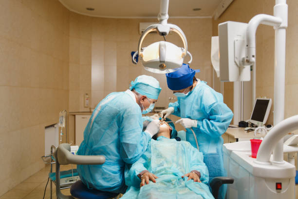 制服と女性アシスタントの男性の古いプロの歯科医師の外科医は、現代のツール装備クリニック光オフィスで女性患者の操作インストール歯科インプラント歯を実行するのに役立ちます。 - dental light dental equipment hospital professional occupation ストックフォトと画像