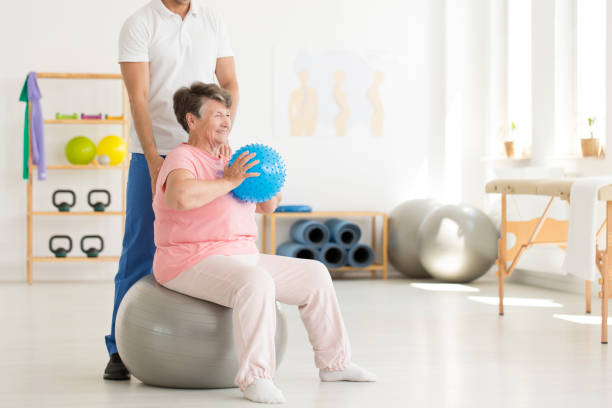 Senior woman exercising in clinic Happy senior woman sitting on grey ball and holding blue ball while exercising at gym parkinsons disease photos stock pictures, royalty-free photos & images