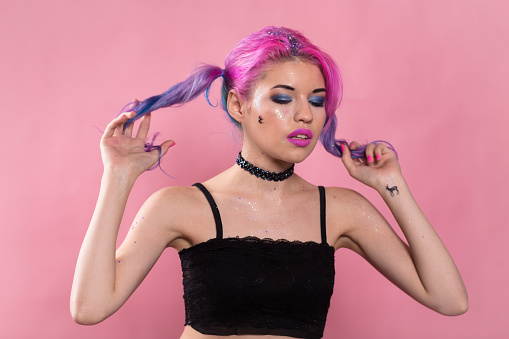 Portrait of heavily madeup woman in black top and choker holding her tails in arms and looking away. Little cat sticker on her cheek. Studio. Half length. Pink background.