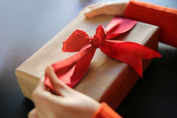 Hand pulls red ribbon on a gift wrapped in brown paper stock photo