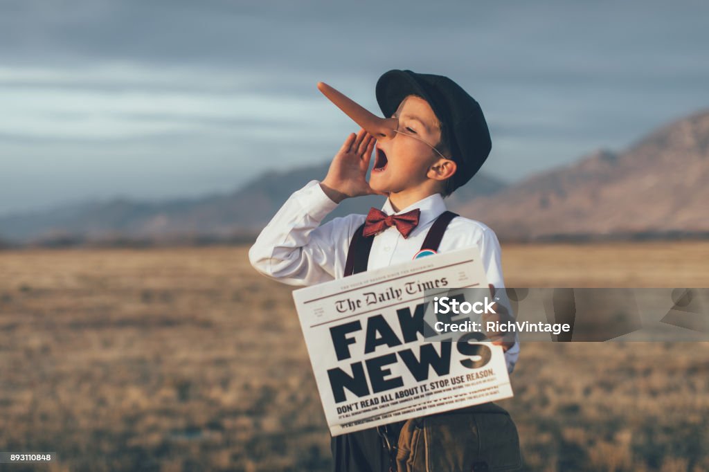 Old Fashioned Pinocchio News Boy Holding Fake Newspaper A news boy dressed in vintage knickers, newsboy hat and fake long Pinocchio nose stands with a fake newspaper in the middle of a field in Utah, USA. He is trying to sell you fake news. Fake News Stock Photo