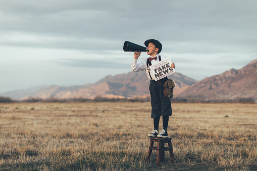 A news boy dressed in vintage knickers and newsboy hat stands with a megaphone and a fake newspaper in the middle of a field in Utah, USA. He yelling loudly trying to sell you fake news.
