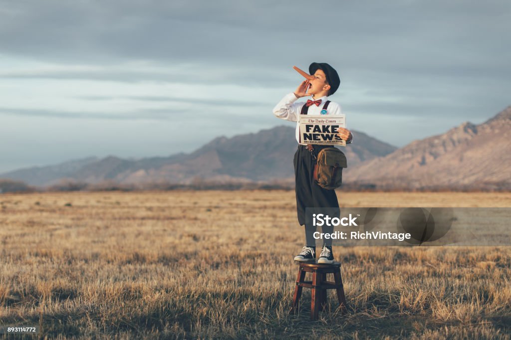 Old Fashioned Pinocchio News Boy Holding Fake Newspaper A news boy dressed in vintage knickers, newsboy hat and fake long Pinocchio nose stands with a fake newspaper in the middle of a field in Utah, USA. He is trying to sell you fake news. Pinocchio Stock Photo