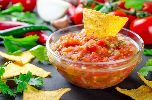 Traditional mexican homemade salsa sauce with ingredients, tomatoes, pepper, cilantro, chips on a dark black stone table. Close-up, horizontal image