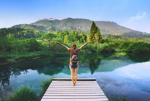 Young woman stands on a wooden bridge with raised arms up on the nature background.