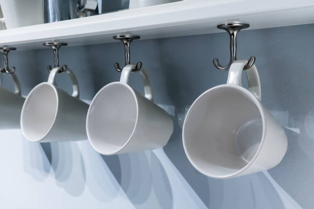 istock Row of white ceramic cups hanging on silver metal hook under white cabinet. Modern interior design. 893259772