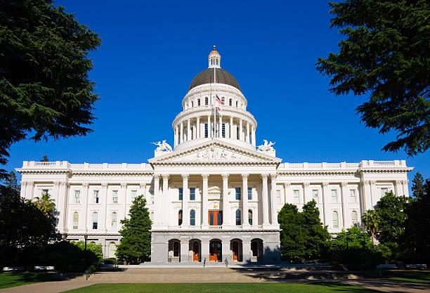 View of the California State Capitol Building under a partly cloudy sky, featuring its iconic white facade, gold dome, and classical columns. The foreground showcases a well-maintained lawn with a few people walking, surrounded by trees and pathways, highlighting a blend of history and public space.