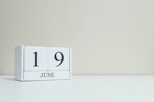 Closeup white wooden calendar with black 19 june word on blurred white wood desk and cream color wallpaper in room textured background with copy space , selective focus at the calendar