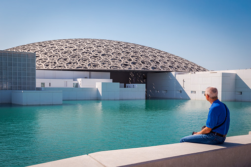 Man in a blue shirt, sitting and looking across a pool of water at the newly opened Louvre, Abu Dhabi, United Arab Emirates. Image taken on November 14, 2017.