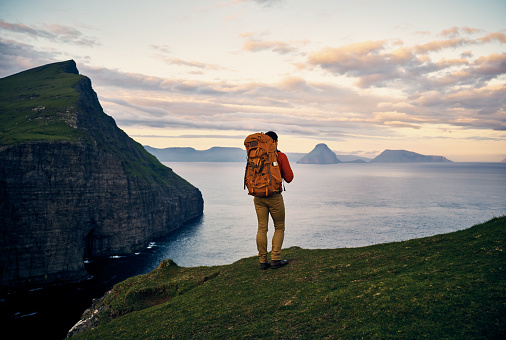 Rearview shot of a unrecognizable man at the edge of a cliff wearing a backpack while looking at the view of the ocean
