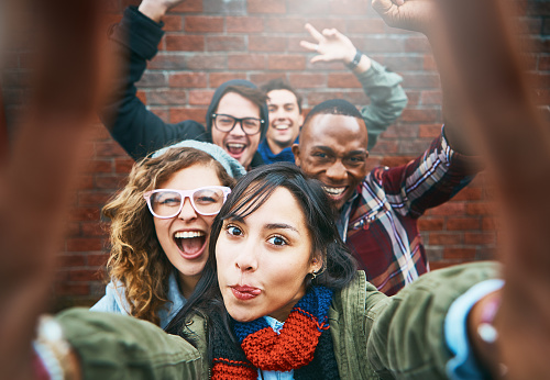 An excited group of young friends waves and laughs, fooling around as they take a selfie outdoors by a brick wall.