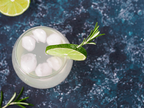Glass of alcoholic coctail with fresh rosemary and lime on blue background. Top view or flat-lay.