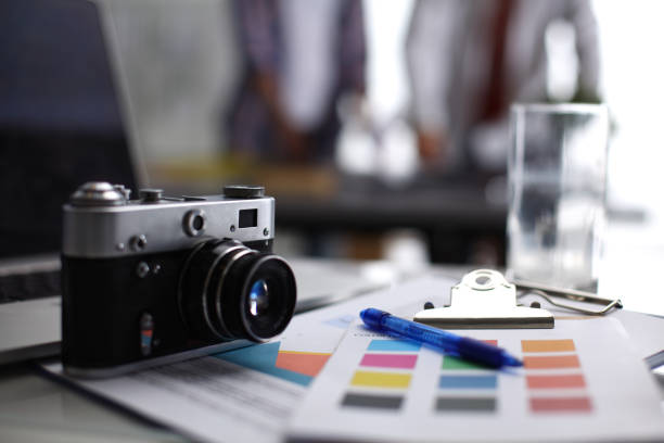 istock Laptop and camera on the desk, two businesspeople standing in the background 893407724