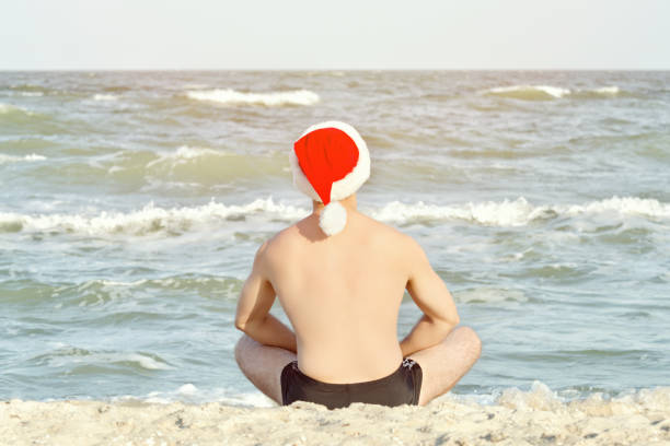 istock Man in Santa hats sits on the sea beach. Back view 893430906