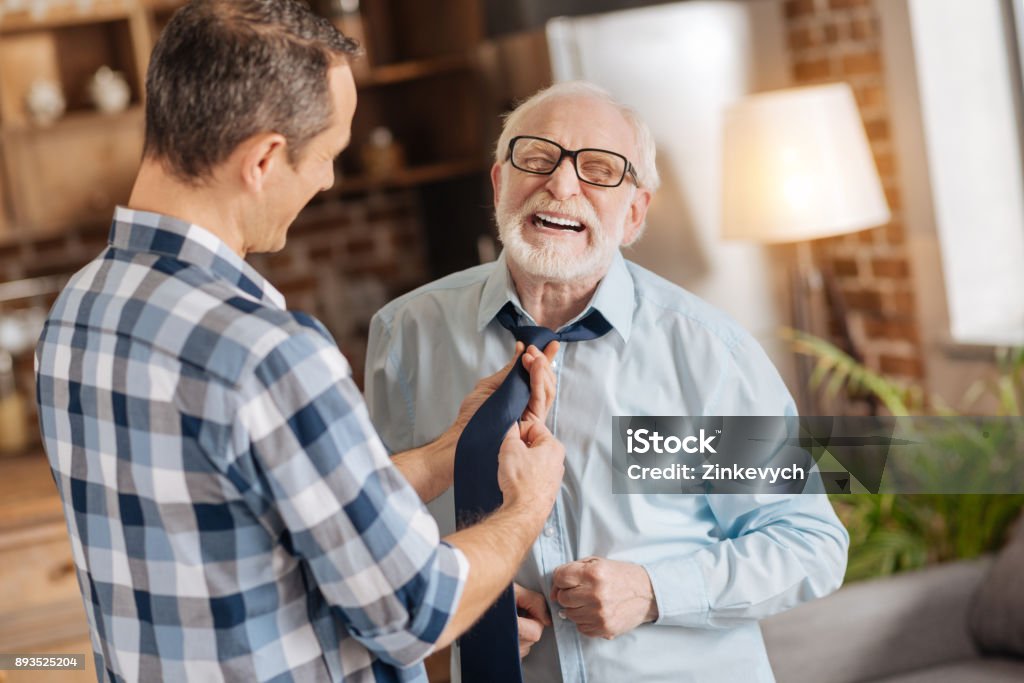 Young man tying the tie of his elderly father Sonly care. Pleasant young man tying the tie of his elderly father while the man laughing, closing his eyes Father Stock Photo