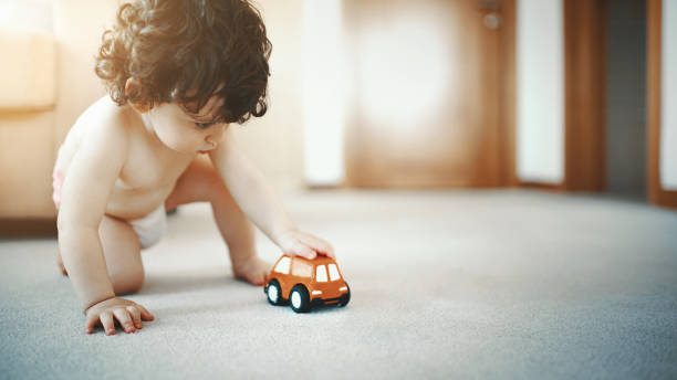 Baby boy playing with a toy car. Closeup of a two year old toddler with curly brown hair sitting on the living room floor and playing with an orange toy car.He's wearing diapers only. first step stock pictures, royalty-free photos & images