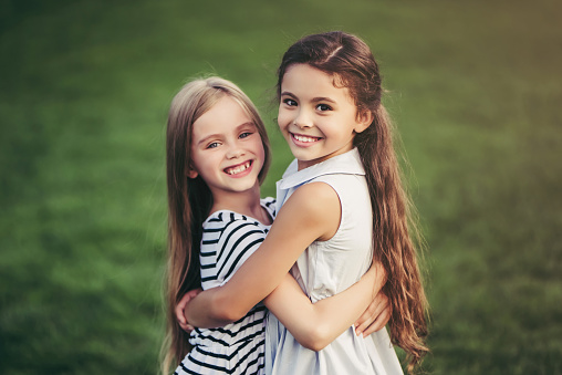Little pretty girls having fun outdoor. Two cute girls are standing on green grass and hugging. Best friends.