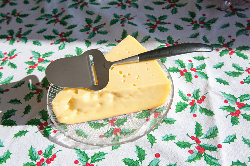 This photo shows a plate with a fresh wedge of cheese on it with a cheese slicer on top of that. The yellow cheese is of a swiss cheese type. The table is brightly lit and covered with a festive Christmas tablecloth with a holly and berry design. A nice and healthy eating food shot with holiday motives. There is a strong shadow that is cast on the green and white tablecloth.- A great shot for preparation cookbooks, ingredient, catalogs, chef, diet and cooking websites or magazines.