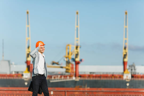 istock Handsome young unshaven successful business man in gray suit and protective construction orange helmet holding case, walking in sea port against cargo ship and crane background in summer time. 893847332