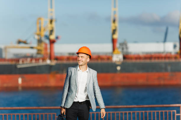 istock Handsome young unshaven successful business man in gray suit and protective construction orange helmet holding case, walking in sea port against cargo ship and crane background in summer time. 893847494