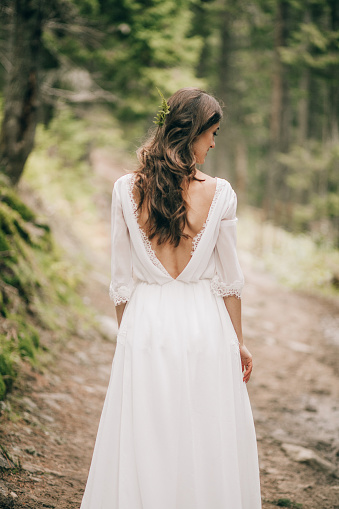 Beautiful bride in whihe dress stands on forest road in Carpathian mountains. Back view.