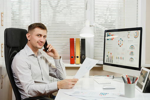 Smiling business man in shirt sitting at the desk, talking on mobile phone, conducting pleasant conversation, working at computer with modern monitor, documents in light office on window background