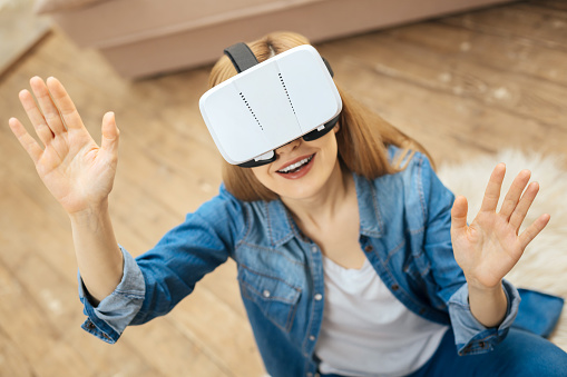 Having fun. Exuberant blond young long-haired woman smiling and wearing virtual reality glasses while sitting on the carpet and wearing a jeans costume