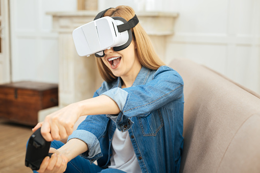 Avid gamer. Cheerful blond young long-haired woman laughing and wearing virtual reality device while sitting on the couch and wearing a jeans jacket and using remote control