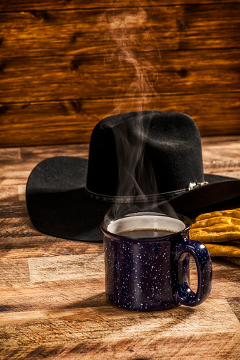 A Blue speckled cup of steaming hot coffee sitting on a rustic wooden table with a black Cowboy hat and leather work gloves on a cold Winter morning at sunrise