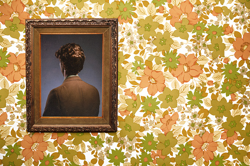 A picture frame hangs on a wall with an ornate frame and retro style, the person posing in the portrait facing the wrong direction, only the back of their head and body visible. Floral wallpaper background.  Horizontal with copy space.