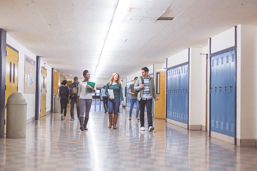 A group of students walk down the hallway between periods. They are talking and smiling and carrying binders, backpacks, and books.