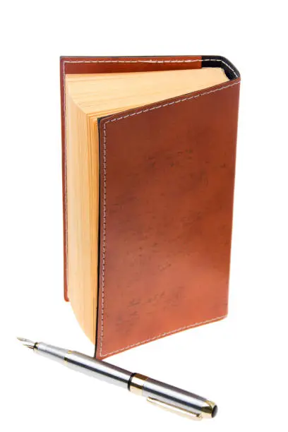 Old brown leather covered book standing on end with a silver fountain pen in front. White background vertical orientation.