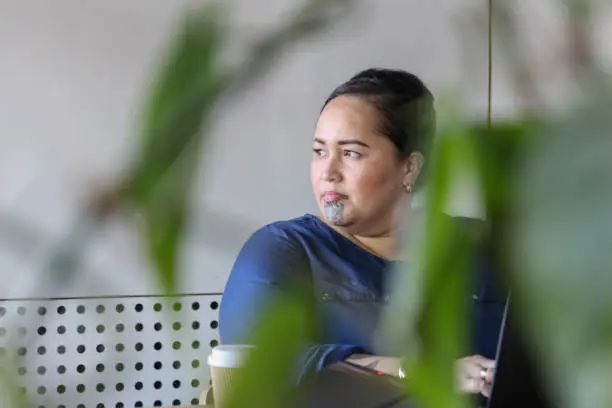 A Maori lady with a moko (facial tattoo), viewed through office foliage, looks to the right of the picture