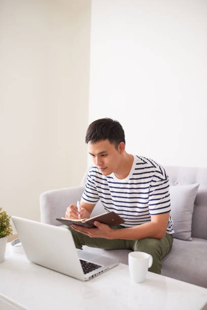 istock Handsome serious man, looking at laptop screen and writing something in his notebook, sitting on couch 894166166