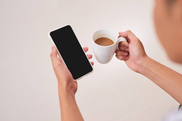 istock Mockup image of woman's hands holding white mobile phone with blank black screen while drinking coffee in modern loft cafe 894172740