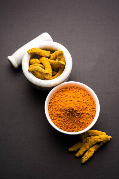 Turmeric powder in ceramic bowl with raw dried turmeric over plain background