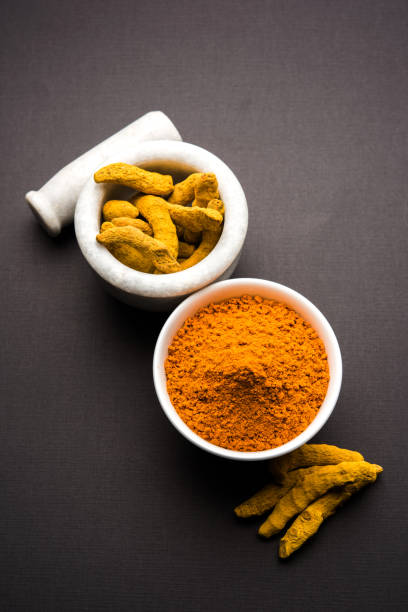 Turmeric powder in ceramic bowl with raw dried turmeric over plain background stock photo