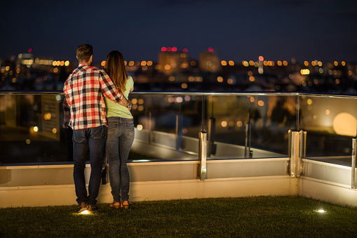 Back view of embraced couple standing on a penthouse balcony at night and looking at view.