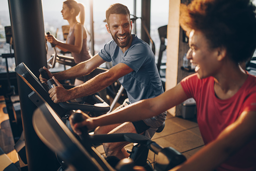 Athletic man exercising on exercise bike in a gym and talking to his female friend next to him.