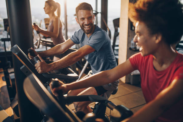 Cheerful male athlete talking to his friend on exercising training in a health club. Athletic man exercising on exercise bike in a gym and talking to his female friend next to him. gym photos stock pictures, royalty-free photos & images