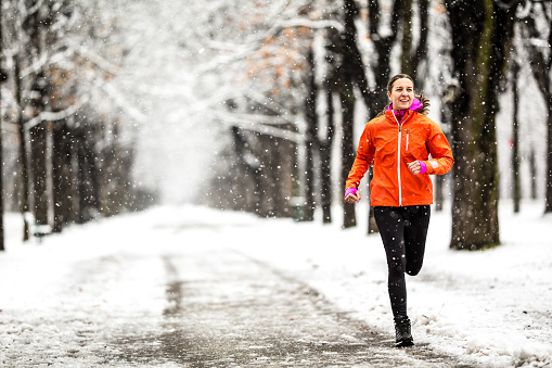 front view of sporty woman jogging  in winter during snowfall on snow covered road in alley with snowcovered trees december january outdoor sports in cold bad weather theme