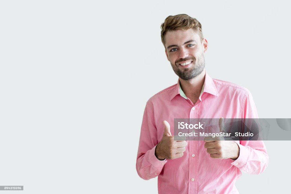 Smiling Young Handsome Man Showing Thumbs Up Closeup portrait of smiling young handsome man looking at camera and showing thumbs up. Isolated front view on white background. Adult Stock Photo